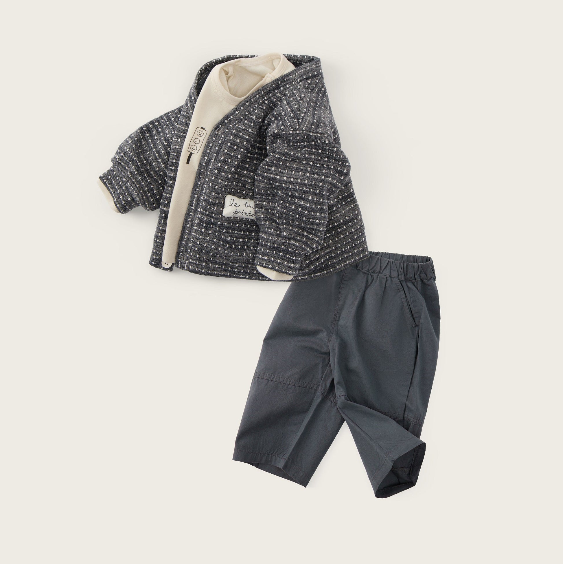 A baby clothing set featuring a dark gray patterned zip-up jacket with a cream lining and matching dark gray pants, displayed on a light beige background with 'jacinda bebe' and 'lovely kids' text.