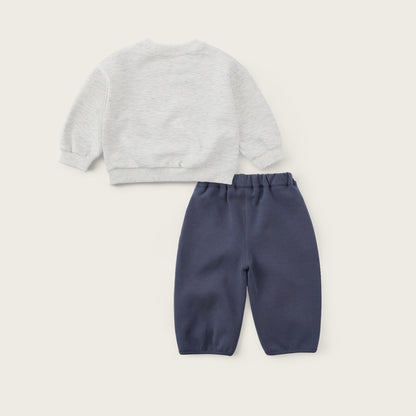 A baby clothing set featuring a light gray heather long-sleeve top and navy blue pants, displayed as a flat lay on a warm off-white background with 'jacinda bebe' and 'lovely kids' text.