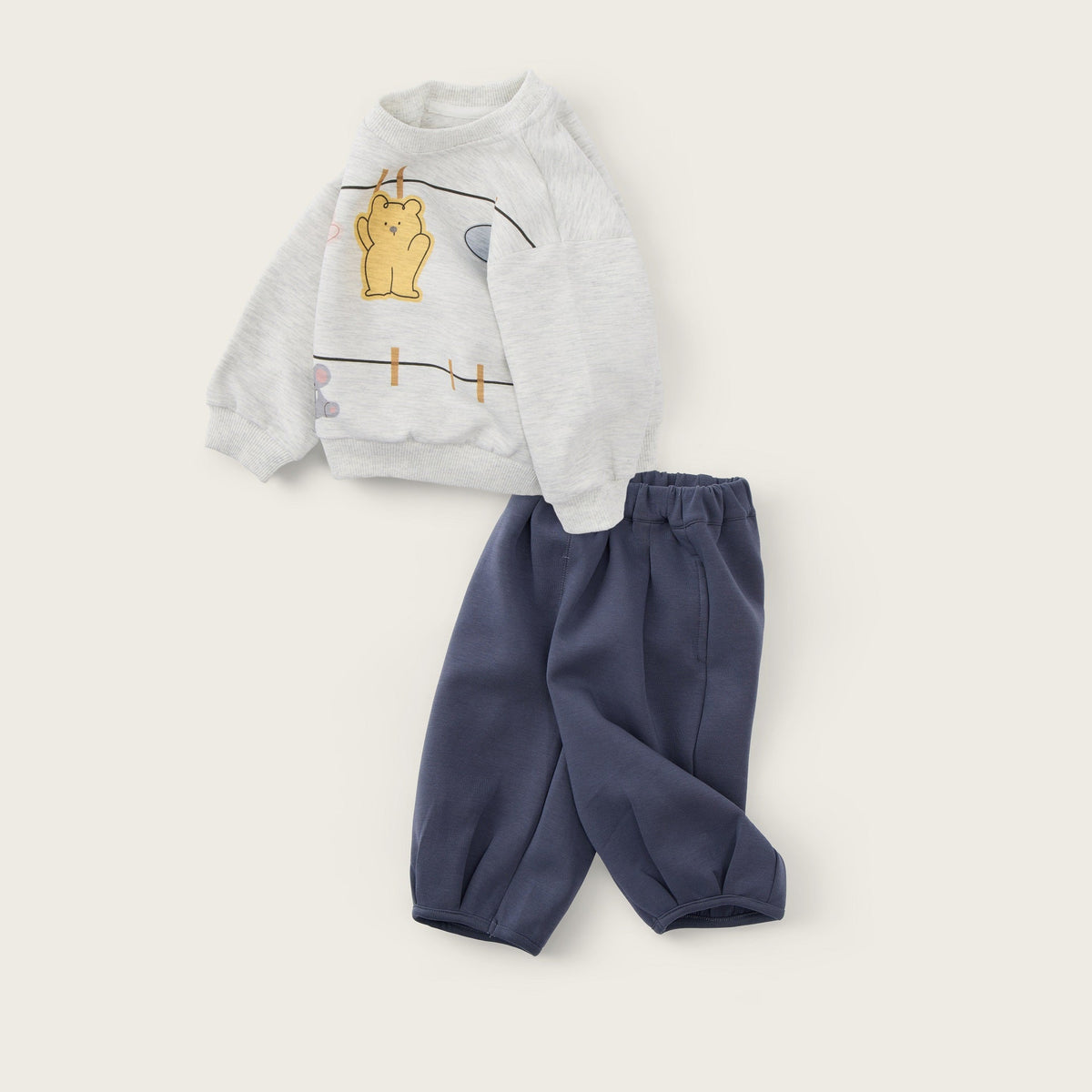 A baby clothing set featuring a light gray sweatshirt with a yellow bear graphic and navy blue pants, displayed on a warm beige background with 'jacinda bebe' and 'lovely kids' text.