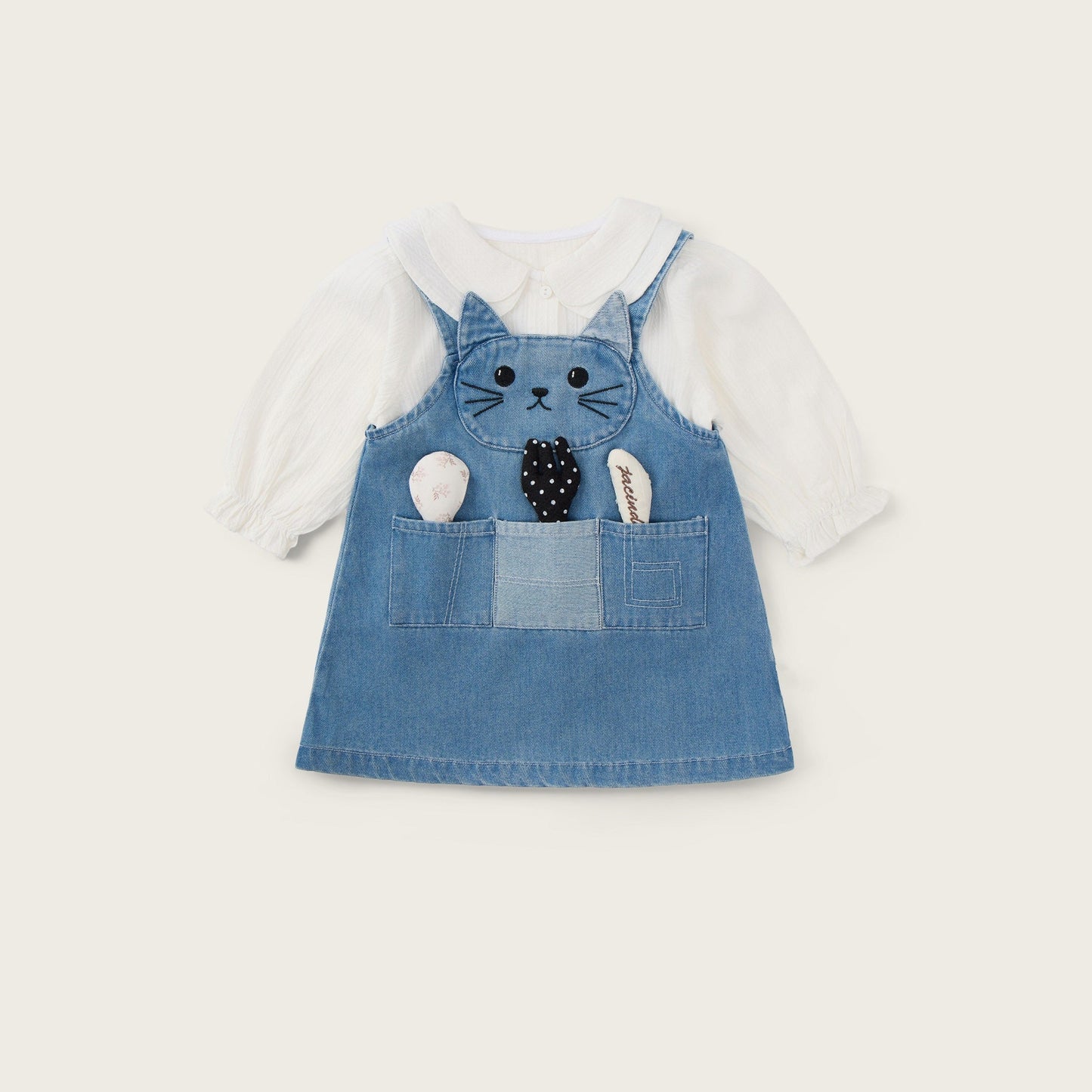 A baby girl's denim overall dress featuring a cat face applique on the chest and a front pocket with plush toy details, layered over a white long-sleeve shirt. The image is presented on a warm cream background with 'jacinda bebe' at the top and 'lovely kids' at the bottom.