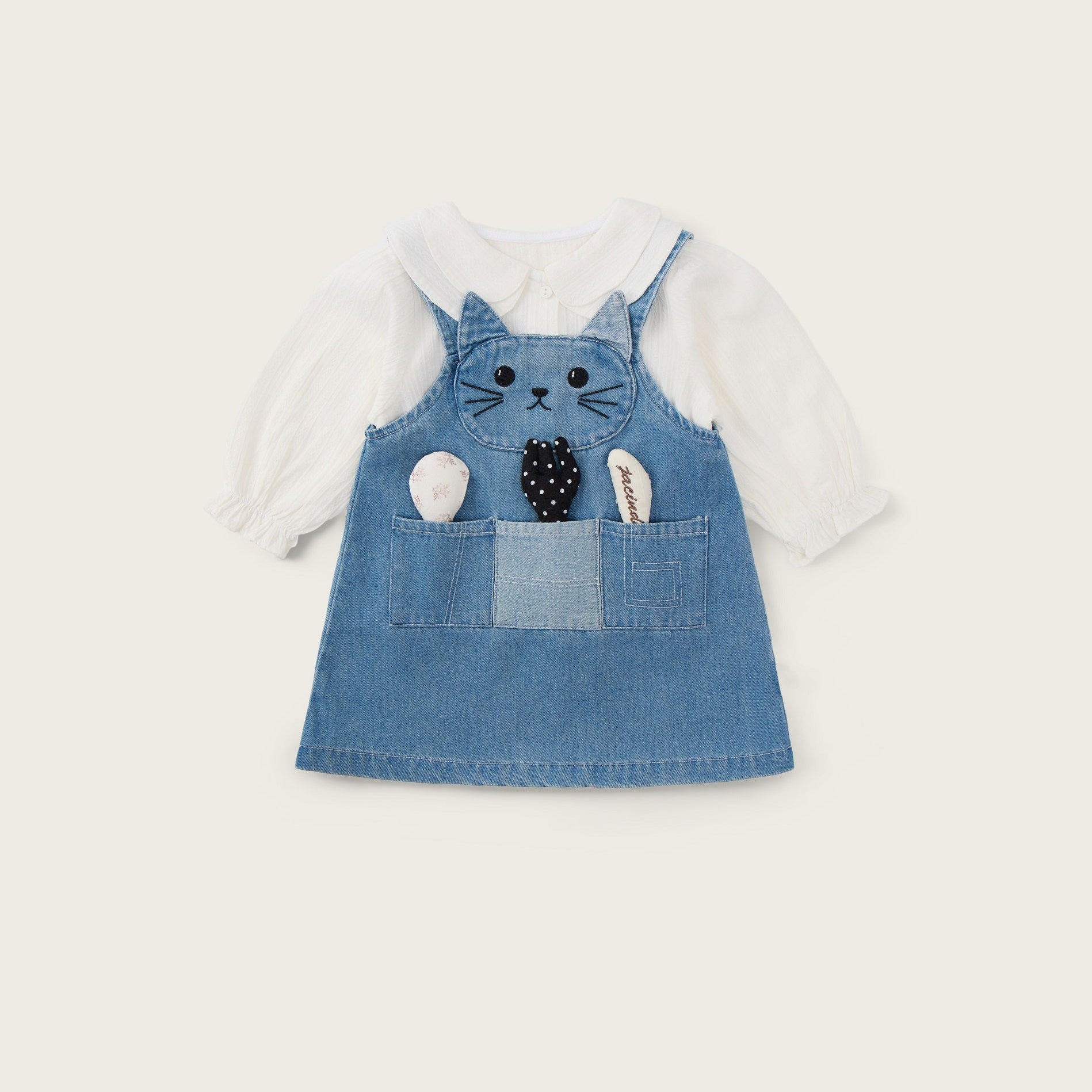 A baby girl's denim overall dress featuring a cat face applique on the chest and a front pocket with plush toy details, layered over a white long-sleeve shirt. The image is presented on a warm cream background with 'jacinda bebe' at the top and 'lovely kids' at the bottom.