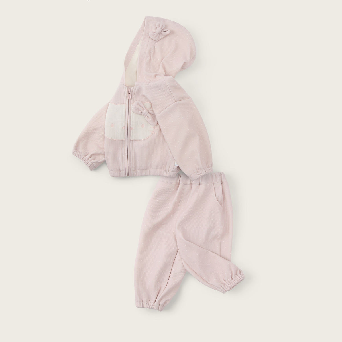 A baby girl's two-piece tracksuit in light pink, featuring a zip-up hoodie with a front patch pocket and matching jogger pants, displayed on a warm off-white background with 'jacinda bebe' and 'lovely kids' text.