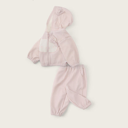 A baby girl's two-piece tracksuit in light pink, featuring a zip-up hoodie with a front patch pocket and matching jogger pants, displayed on a warm off-white background with 'jacinda bebe' and 'lovely kids' text.