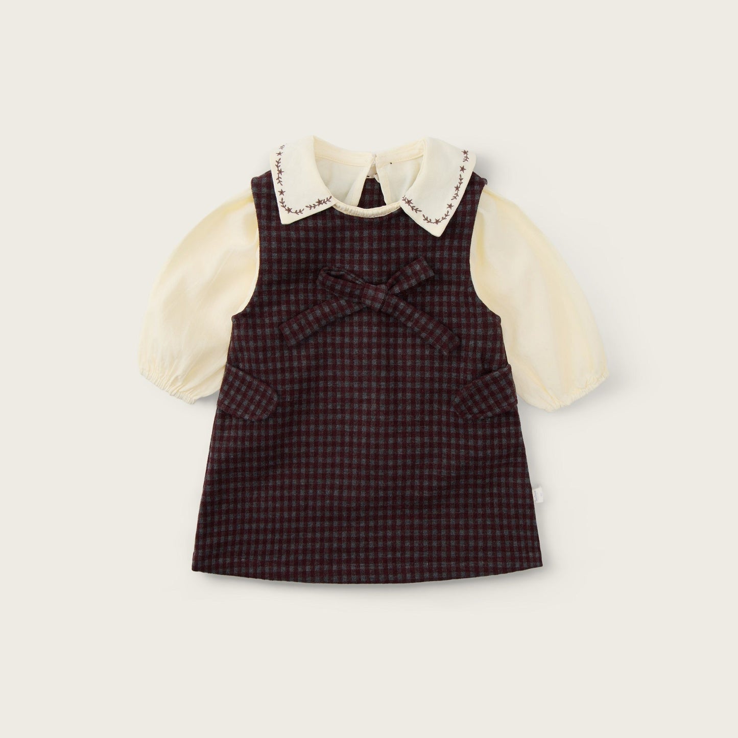 A baby girl's dress featuring a dark brown and red checkered pinafore vest over a cream-colored long-sleeve blouse with a lace-trimmed Peter Pan collar. The brand names 'jacinda bebe' at the top and 'lovely kids' at the bottom are printed on a light beige background.