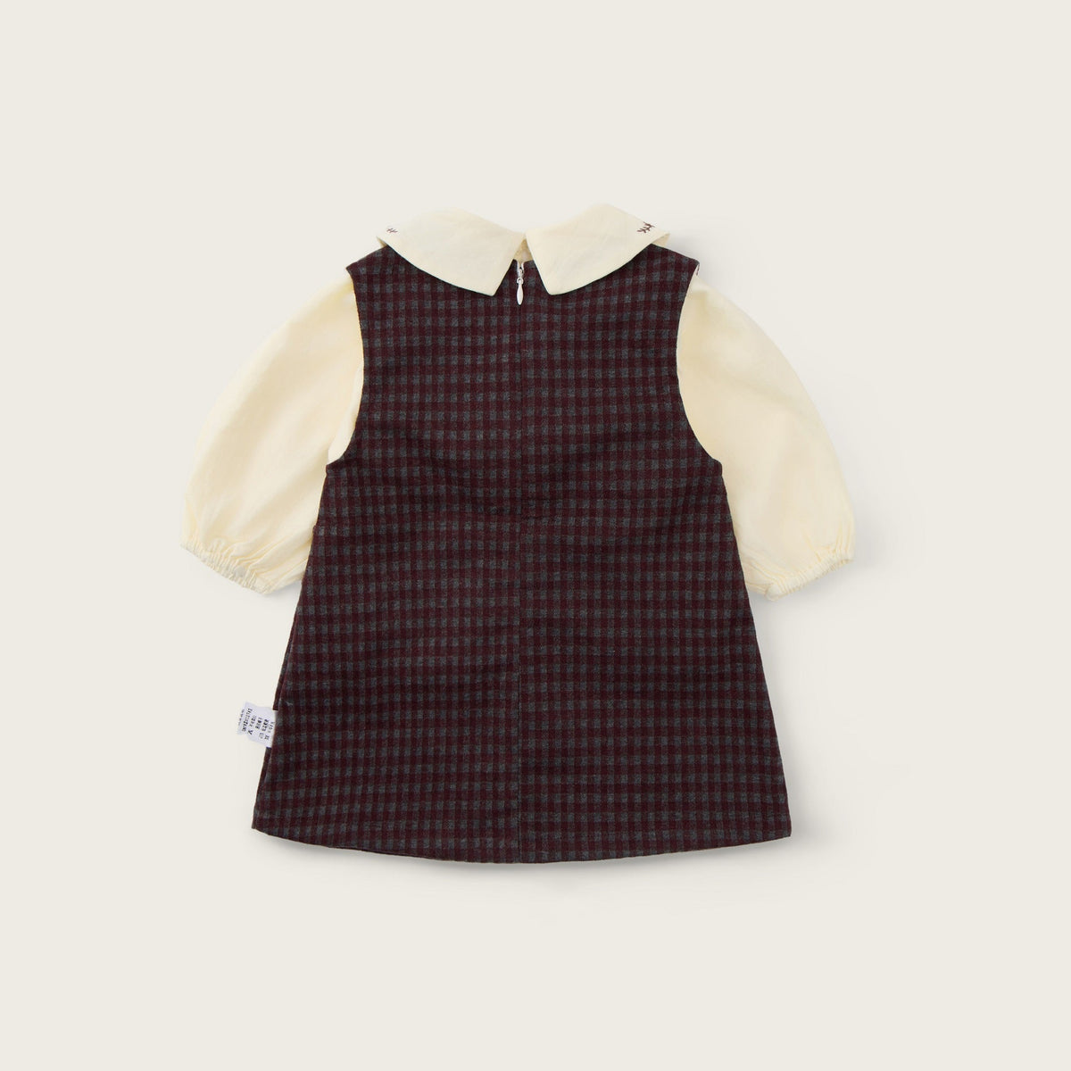 A baby girl's dress by Jacinda Bebe from Lovely Kids, featuring a dark brown and red checkered vest over a cream-colored long-sleeve shirt with a Peter Pan collar, displayed on a light beige background with brand text.