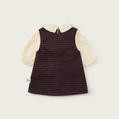 A baby girl's dress by Jacinda Bebe from Lovely Kids, featuring a dark brown and red checkered vest over a cream-colored long-sleeve shirt with a Peter Pan collar, displayed on a light beige background with brand text.