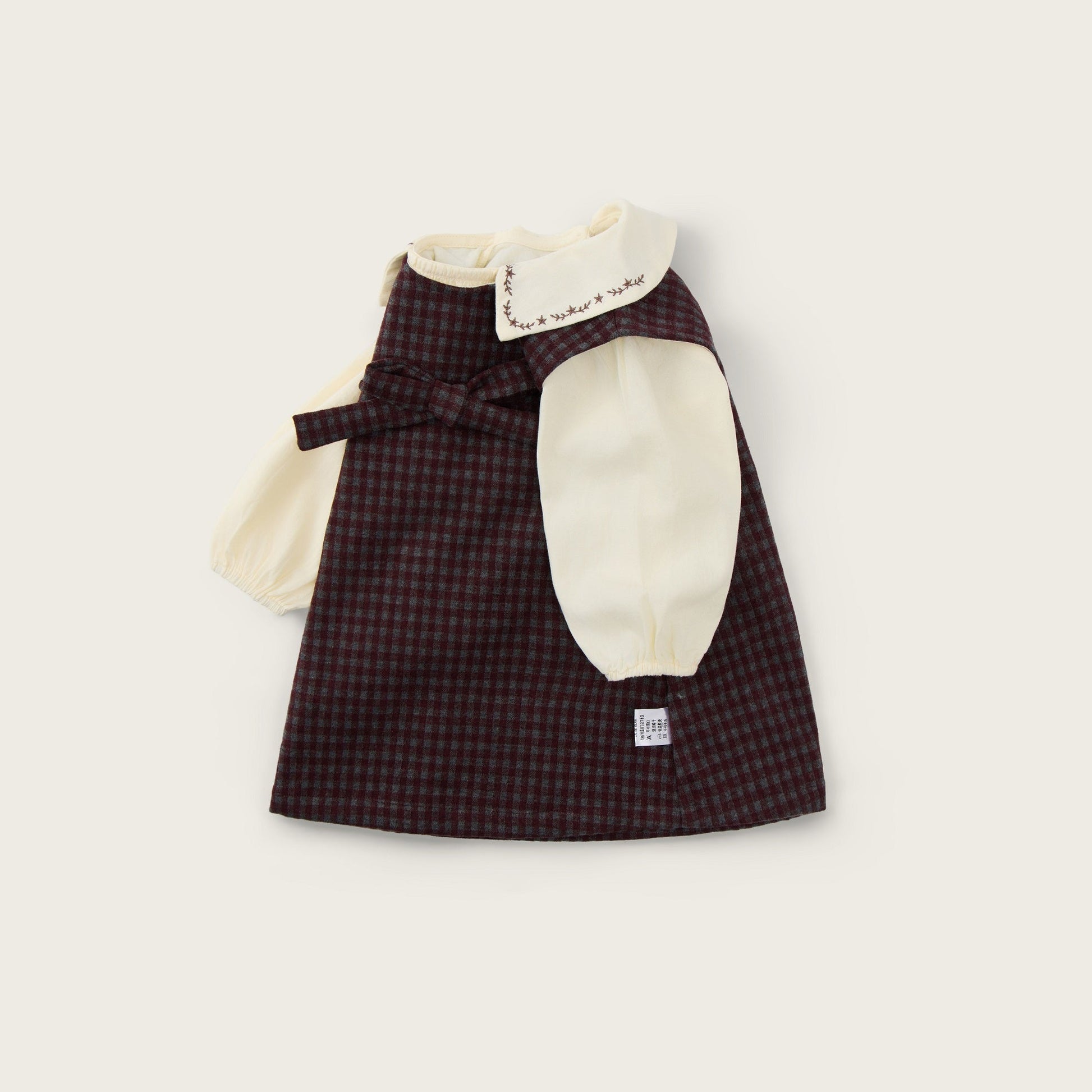 A baby girl's dress from Jacinda Bebe, featuring a dark brown and black checkered skirt, cream puff sleeves, and a cream collar with embroidered details. The dress is displayed flat on a light beige background with 'jacinda bebe' written at the top and 'lovely kids' at the bottom.