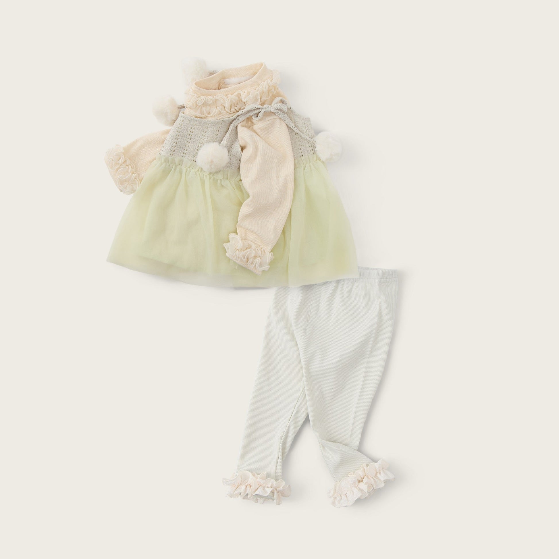 A baby girl's two-piece outfit set featuring a pale yellow-green tulle dress with a cream knit top and pom-pom details, paired with cream leggings with ruffle trim, displayed on a light beige background with 'jacinda bebe' and 'lovely kids' text.