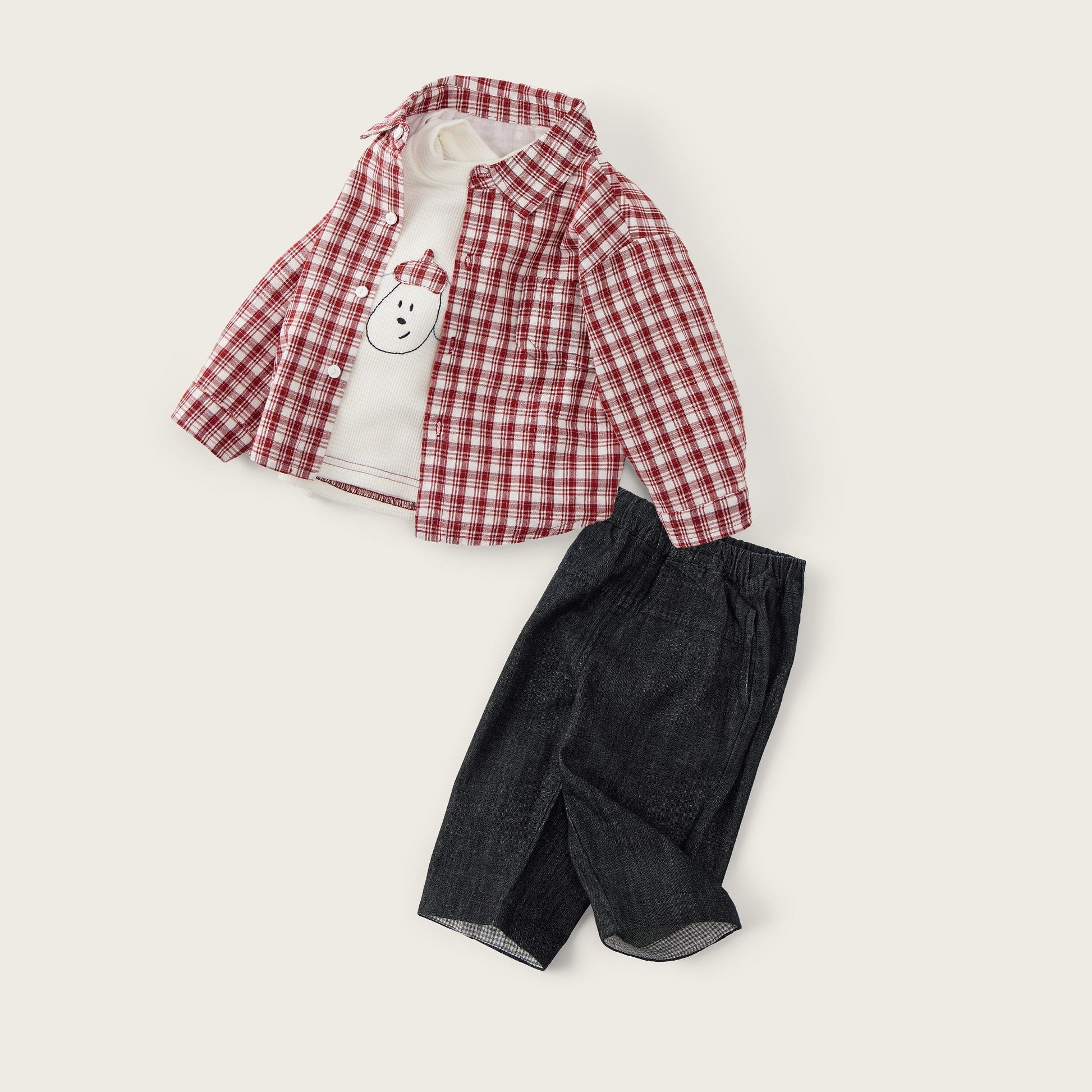 A children's outfit set laid flat on a light cream background, featuring a red and white plaid button-up shirt over a white long-sleeve top with a small bear face patch, paired with dark charcoal shorts. The text 'jacinda bebe' appears at the top and 'lovely kids' at the bottom.