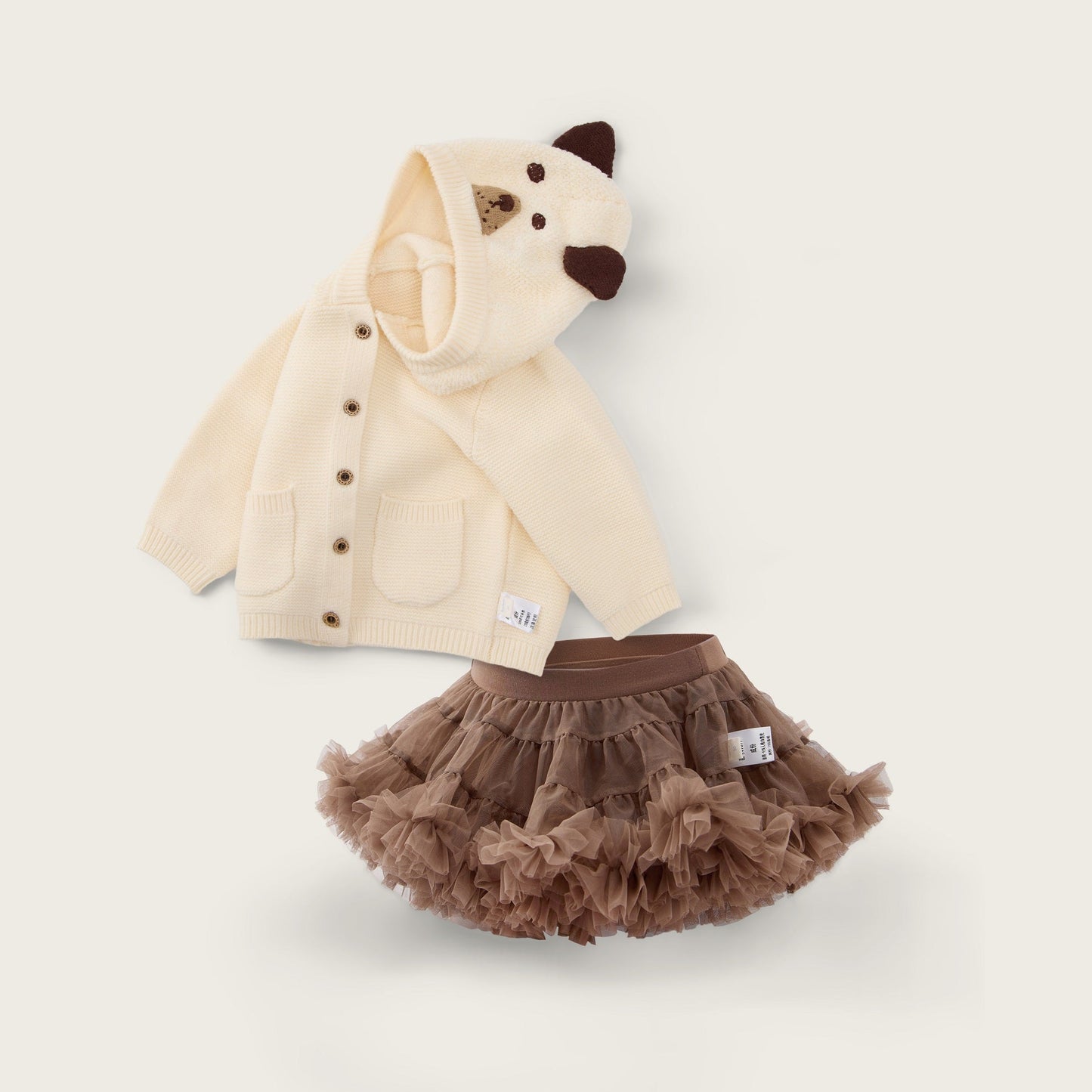 A baby clothing set featuring a cream-colored hooded cardigan with a bear face on the hood and brown buttons, paired with a brown ruffled tutu skirt, displayed on a light beige background with decorative text.